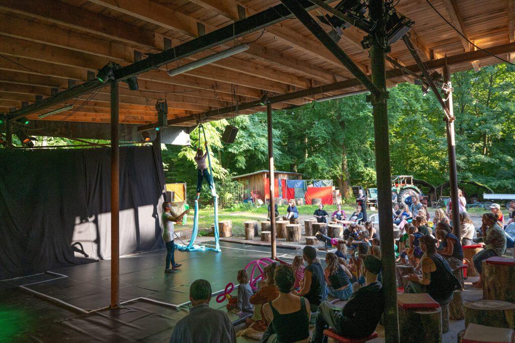 Ein Freilufttheater mit Holzüberdachung, in dem eine Artistin an einem blauen Vertikaltuch schwebt, während eine andere Person sie sichert. Im Publikum sitzen Familien und Kinder auf Baumstämmen und Bänken und verfolgen die Aufführung aufmerksam. Im Hintergrund ist grüner Wald und ein kleines Holzhaus zu sehen.