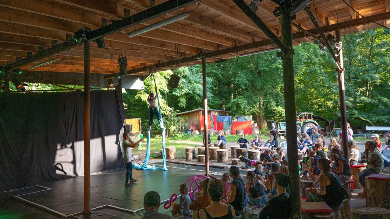 Eine Artistin führt auf einer überdachten Freiluftbühne eine Nummer am blauen Vertikaltuch auf. Sie hängt in der Luft, während eine zweite Person am Boden das Tuch hält. Vor der Bühne sitzen Kinder und Erwachsene auf Baumstämmen und Holzbänken und verfolgen gespannt die Vorstellung. Im Hintergrund ist dichter grüner Wald und ein kleines Holzhaus zu sehen – die Szene wirkt lebendig und sommerlich.