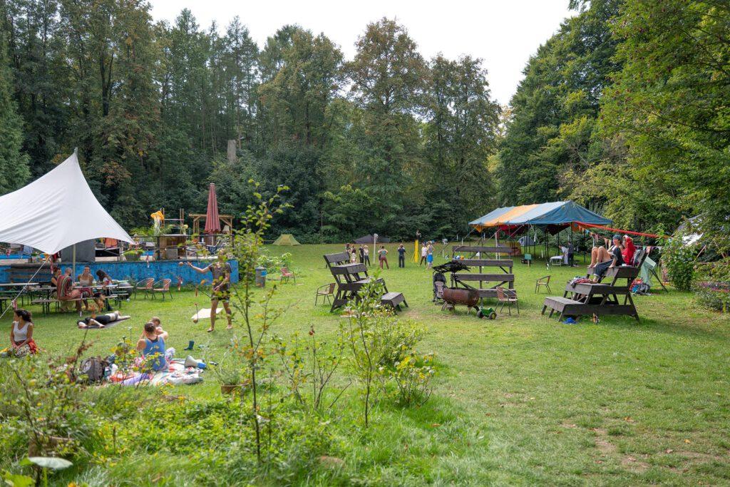 Eine offene Wiese mit Zelten, Bänken und Menschen, die entspannt zusammensitzen oder jonglieren. Einige liegen auf Decken im Gras, andere spielen oder unterhalten sich. Im Hintergrund sind bunte Zelte und dichte Bäume zu sehen, was eine sommerliche Festivalatmosphäre vermittelt.