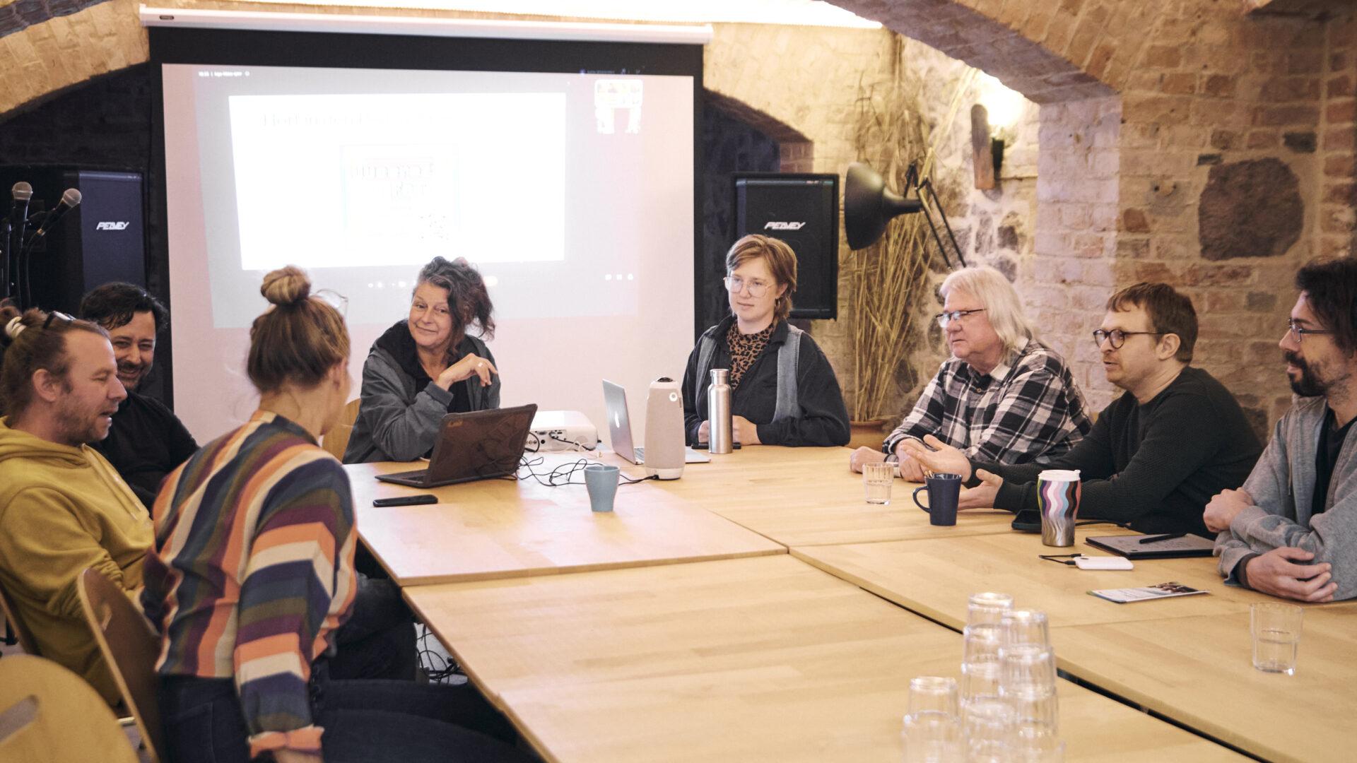 Eine Gruppe von Menschen sitzt an zusammengestellten Holztischen in einem Gewölbekeller und unterhält sich; im Hintergrund eine Leinwand mit einer Präsentation, auf dem Tisch Laptops und eine 360°-Konferenzkamera.